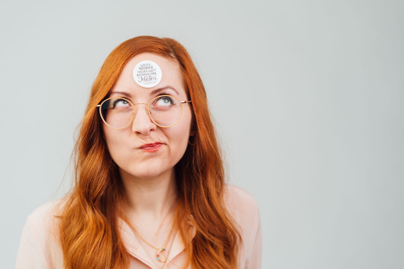 Eine Frau mit langen roten Haaren und Brille blickt mit einem verspielten Ausdruck nach oben. Sie hat einen runden Aufkleber auf der Stirn. Der Hintergrund ist einfarbig und hell.
