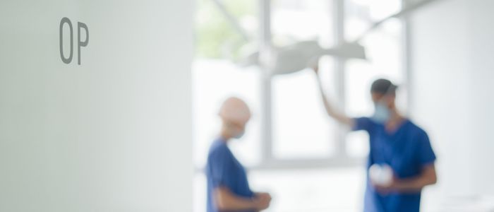 Unscharfes Bild von zwei Chirurgen in blauen Kitteln und Masken, die über einem Patienten in einem Operationssaal stehen, mit der Aufschrift OP auf einer Glaswand im Vordergrund.