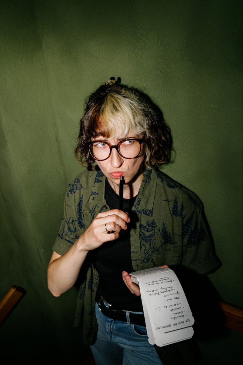 Eine Person mit Brille und lockigem Haar steht vor einer grünen Wand und hält ein Notizbuch und einen Stift in der Hand. Sie scheint nachzudenken, denn der Stift berührt ihre Lippen und sie hat einen nachdenklichen Ausdruck im Gesicht.