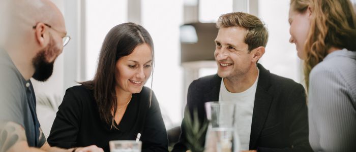 Vier Personen sitzen an einem Tisch in einem hellen Büro, lächeln und diskutieren. Auf dem Tisch liegen Papiere, ein Glas Wasser und eine Pflanze. Die Atmosphäre wirkt gemeinschaftlich und freundlich.