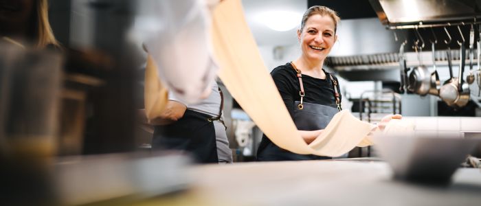Eine lächelnde Frau in einer Schürze dehnt ein langes Stück Nudelteig in einer professionellen Küche aus, während eine weitere Person neben ihr steht und Kochutensilien im Hintergrund hängen.