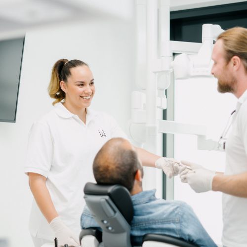 Ein Zahnarzt und eine Zahnarzthelferin, beide in weißen Kitteln und Handschuhen, lächeln und sprechen mit einem sitzenden Patienten in einer modernen, hellen Zahnklinik.