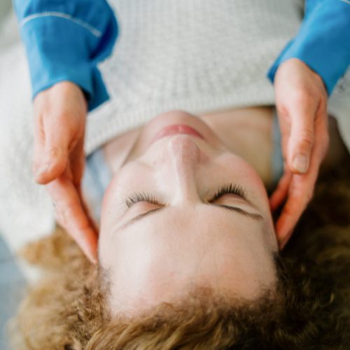 Eine Person mit lockigem Haar liegt mit geschlossenen Augen auf dem Rücken, während eine andere Person ihre Hände sanft an die Seiten des Kopfes legt, was auf eine beruhigende oder therapeutische Behandlung hindeutet.