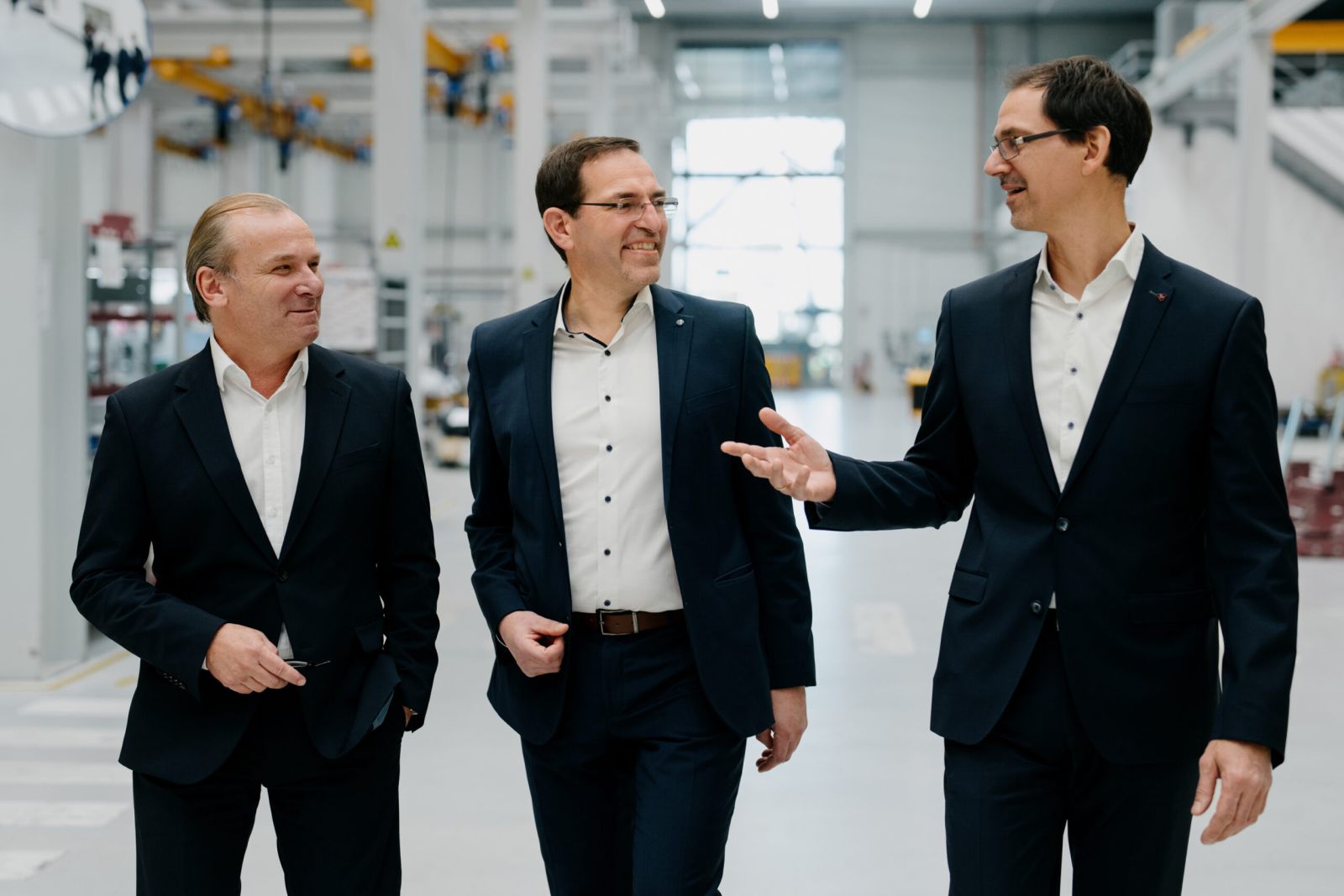 Drei Männer in Geschäftsanzügen gehen in einem geräumigen, modernen Industriegebäude mit hohen Decken und großen Fenstern zusammen, lächeln und unterhalten sich.