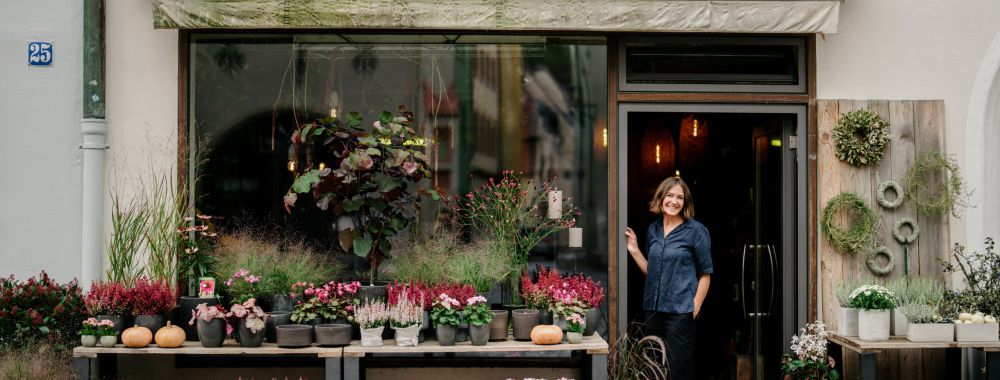 Eine Frau steht lächelnd in der Tür eines Blumenladens, der sich "Blumenladen" nennt, umgeben von verschiedenen Topfpflanzen und Blumen, die in Holzkisten auf dem Bürgersteig stehen.