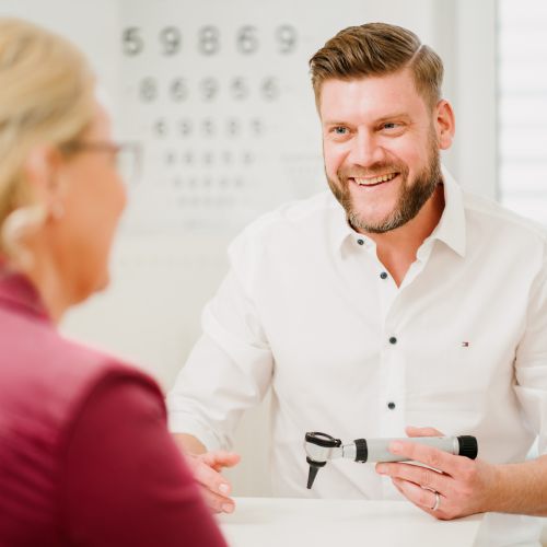 Ein lächelnder Optometrist im weißen Hemd hält ein Ophthalmoskop in der Hand, während er sich mit einer Patientin in einer roten Jacke unterhält, während im Hintergrund eine Augentabelle zu sehen ist.