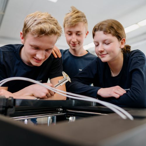 Drei Jugendliche in marineblauen Hemden untersuchen einen 3D-Drucker, wobei einer einen Messschieber hält, um ein gedrucktes Objekt zu messen, während die anderen in einem hellen Klassenzimmer aufmerksam zusehen.