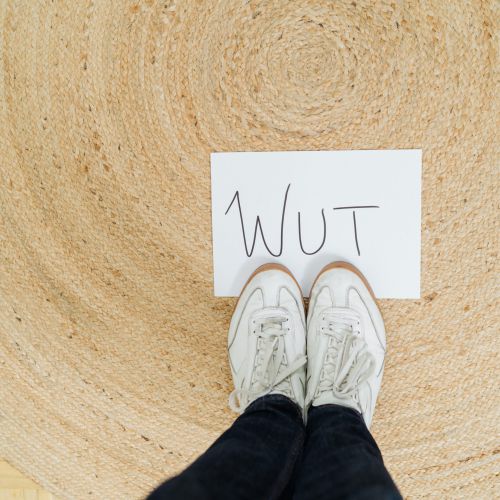 Eine Person in weißen Turnschuhen steht auf einem runden Webteppich, vor ihr liegt ein Stück Papier auf dem Boden, auf dem in großen Buchstaben WUT geschrieben steht.