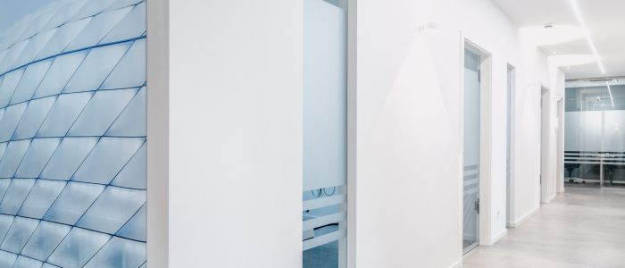 Ein moderner, heller Flur mit weißen Wänden, offenen Türen zu kleinen Räumen und großen Fenstern, die ein geometrisches Muster aus einem angrenzenden Gebäude widerspiegeln. Der Boden ist mit hellgrauen Fliesen ausgelegt, die ein klares, minimalistisches Erscheinungsbild erzeugen.