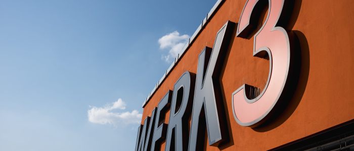 Große, fette Buchstaben und eine Zahl WERK 3 sind auf einer orangefarbenen Gebäudefassade vor einem klaren blauen Himmel mit ein paar kleinen Wolken angebracht. Die Perspektive ist von unten nach oben geneigt.