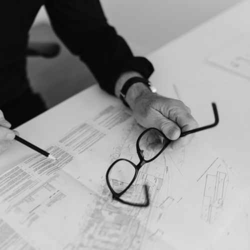 Eine Person in einem dunklen Hemd sitzt an einem Schreibtisch, hält in der einen Hand eine Brille und deutet mit einem Stift in der anderen Hand auf Text und Diagramme in einem Dokument. Das Bild ist in schwarz-weiß gehalten.