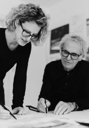 Eine Frau und ein älterer Mann, beide mit Brille, arbeiten an einem Schreibtisch in einem hellen Büro an Skizzen, während hinter ihnen Pläne und Bilder an der Wand hängen. Das Bild ist schwarz-weiß.