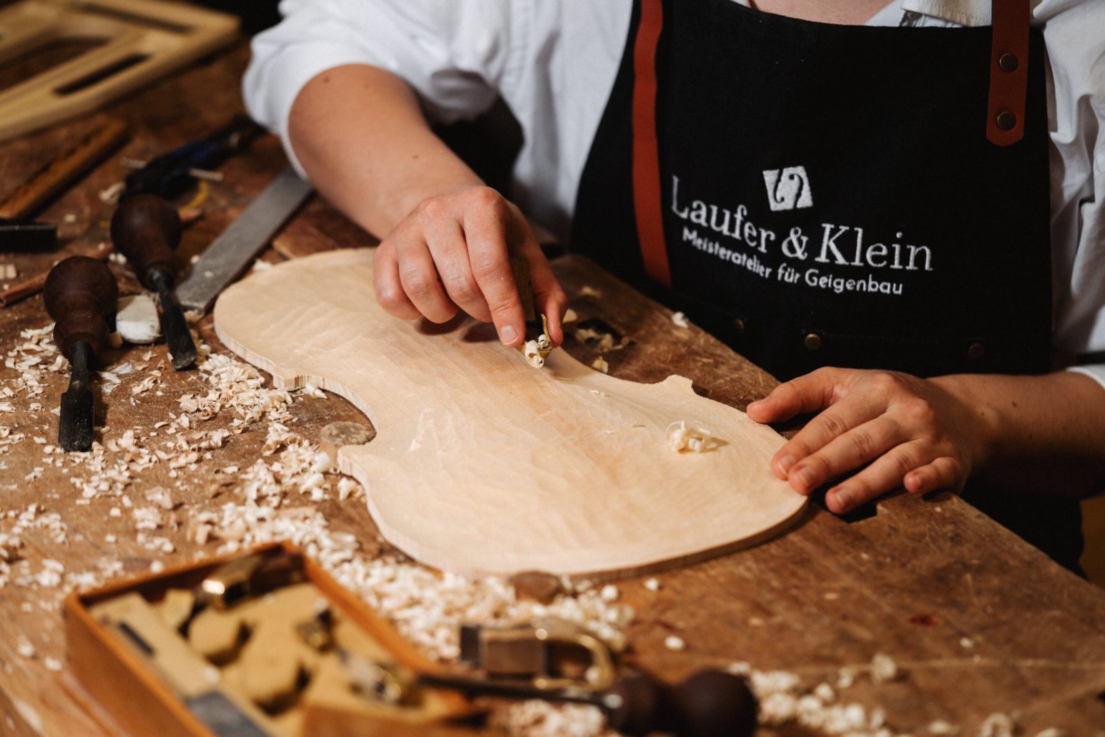 Eine Person mit schwarzer Schürze arbeitet an einer Werkbank, umgeben von Holzspänen und Holzbearbeitungswerkzeugen, an der Schnitzerei einer hölzernen Geigendecke.