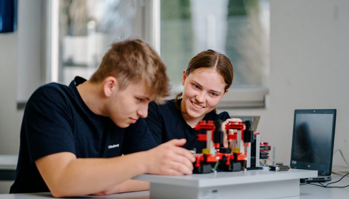 Zwei junge Leute in dunklen Hemden arbeiten gemeinsam an einem mechanischen Modell aus bunten Plastikteilen an einem Tisch. Einer justiert das Modell, während der andere lächelt. In der Nähe ist ein Laptop aufgeklappt.