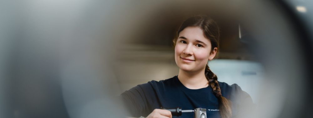Eine junge Frau mit einem Zopf lächelt, während sie ein handgehaltenes Elektrowerkzeug benutzt, gesehen durch einen kreisförmigen, verschwommenen Vordergrund.