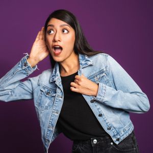 Eine Frau in Jeansjacke und schwarzem Hemd hält sich eine Hand ans Ohr und sieht überrascht aus, während sie vor einem lila Hintergrund steht.
