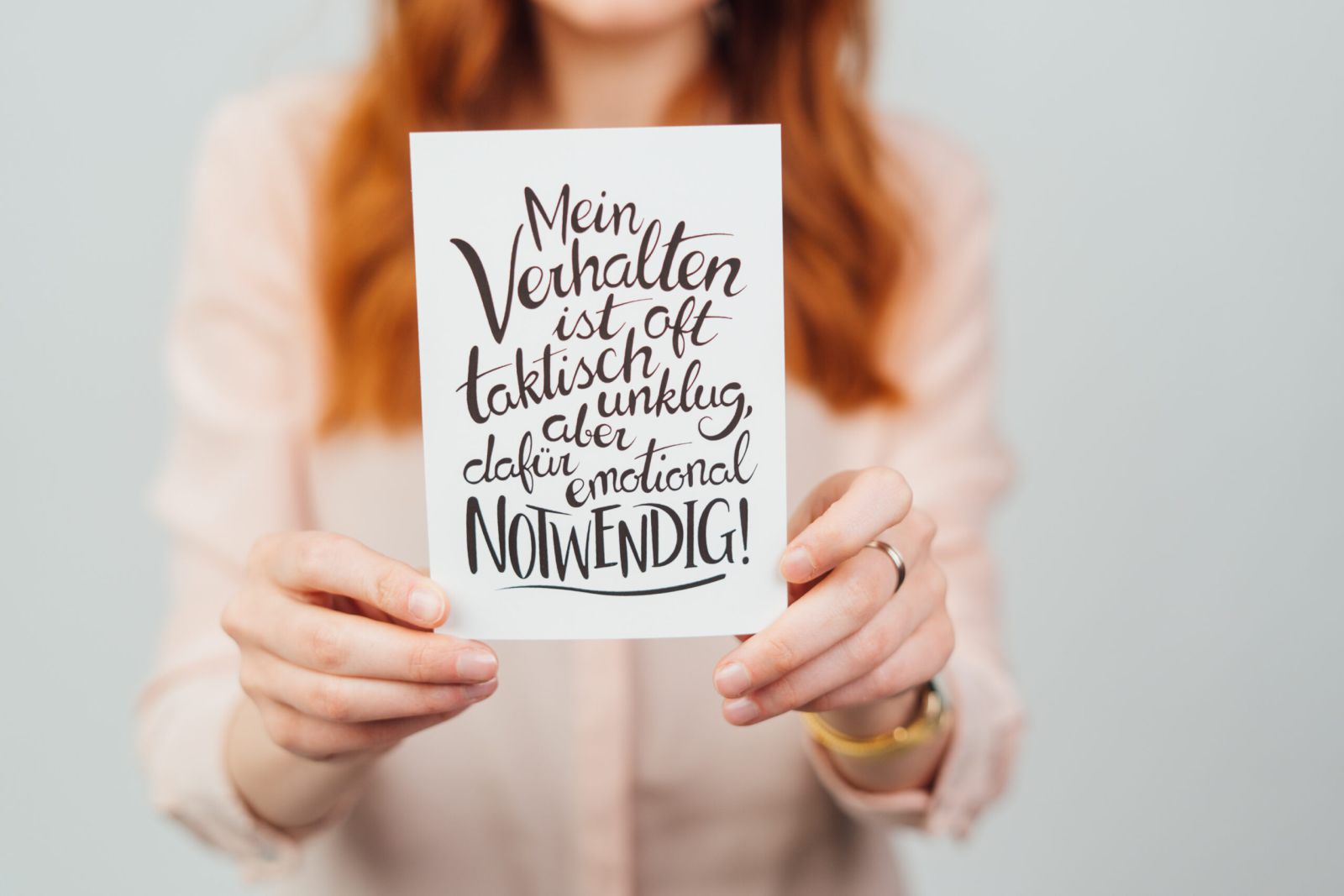 Eine Frau mit langen roten Haaren hält eine Karte mit einem handgeschriebenen deutschen Text, auf dem steht: Mein Verhalten ist oft taktisch unklug, aber dafür emotional notwendig. Der Hintergrund ist hell und unscharf.