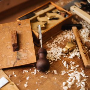 Eine hölzerne Werkbank, die mit Holzspänen bedeckt ist, verschiedene Holzbearbeitungswerkzeuge, darunter ein Meißel mit Holzgriff, ein Hammer, ein Messwerkzeug und eine offene Holzkiste mit Metallteilen.