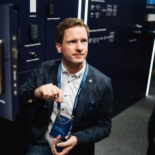 Ein Mann im Anzug kniet neben einem dunkelblauen Türdisplay, hält eine Broschüre in der Hand und blickt nach oben. Das Display zeigt moderne Türgriffe und Sicherheitseinrichtungen in einem gut beleuchteten Ausstellungsraum.