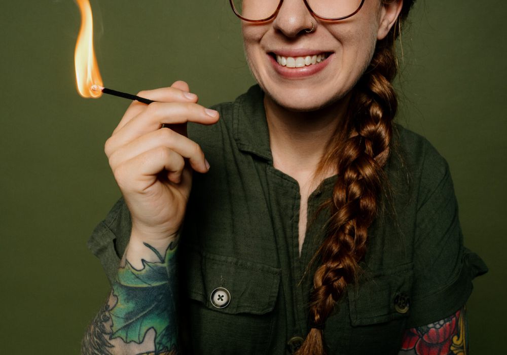 Eine lächelnde Person mit Brille, einem langen roten Zopf und bunten Armtattoos hält ein brennendes Streichholz vor einem olivgrünen Hintergrund.