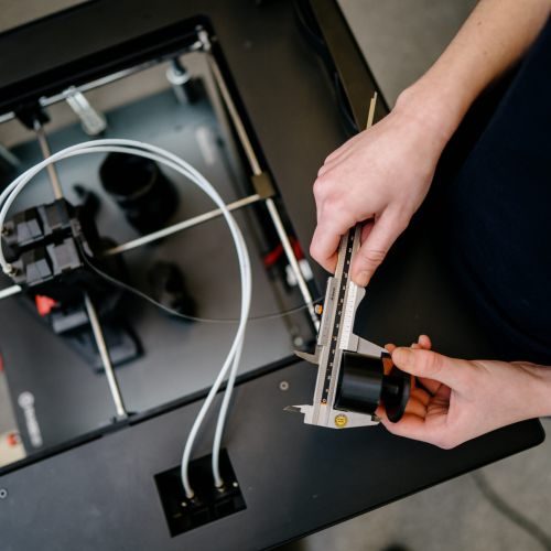 Eine Person misst mit einem Messschieber ein 3D-gedrucktes Objekt neben einem 3D-Drucker, bei dem Drähte und Komponenten sichtbar sind.