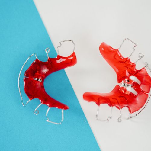 Zwei rote kieferorthopädische Retainer mit Metalldrähten sind auf einem diagonal zwischen Blau und Weiß geteilten Hintergrund platziert.