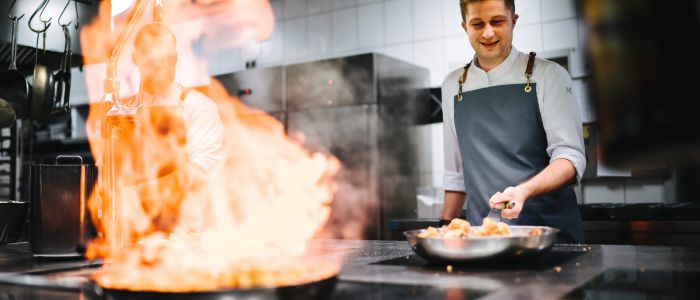 Ein Koch in einer modernen Küche lächelt beim Kochen und hält eine Pfanne, während eine große Flamme aus einer anderen Pfanne auf dem Herd aufsteigt. Die Küche ist sauber und hell erleuchtet, mit Geräten aus Edelstahl und weiß gekachelten Wänden.