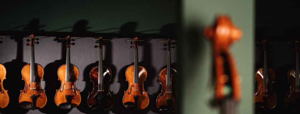 Eine Reihe von Geigen hängt an einer dunklen Wand, die von weichem Licht beleuchtet wird. Im Vordergrund auf der rechten Seite des Bildes ist ein verschwommenes Streichinstrument zu sehen.