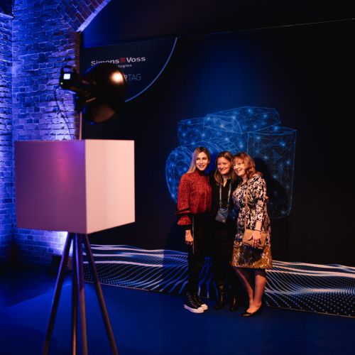Drei Frauen posieren und lächeln gemeinsam vor einem dunklen geometrischen Hintergrund bei einer Veranstaltung. Im Vordergrund befindet sich eine professionelle Beleuchtungsanlage, und die Kulisse besteht aus blauen und violetten Lichtern und freiliegenden Ziegelwänden.