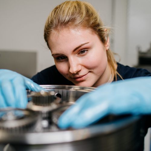 Eine Person mit blauen Handschuhen prüft und justiert Metallzahnräder in einer Maschine und konzentriert sich dabei auf die Aufgabe in einem Labor oder einer industriellen Umgebung.