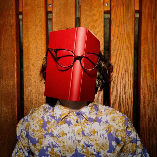 Eine Person, die ein geblümtes Hemd trägt, sitzt auf einer Holzbank und hat ein rotes Buch vor dem Gesicht. Auf dem Buch ist eine Brille angebracht, die den Eindruck erweckt, das Buch habe ein Gesicht.