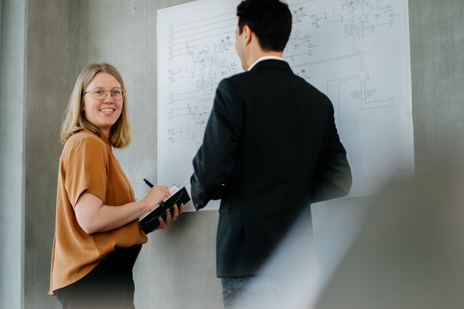 Eine Frau mit einem Notebook lächelt in die Kamera, während sie neben einem Mann im Anzug steht, der ein großes technisches Diagramm an der Wand betrachtet.