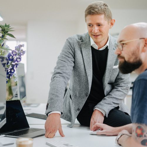 Zwei Männer arbeiten in einem Büro zusammen an einem Schreibtisch mit einem Laptop. Ein Mann in einem grauen Blazer zeigt auf ein Dokument, während der andere, mit Brille und Tattoos, sitzt und auf den Bildschirm schaut. Auf dem Schreibtisch liegen Blumen und Büromaterial.