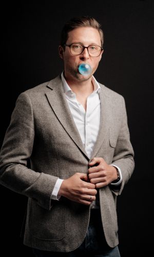 Ein Mann mit Brille, grauem Blazer und weißem Hemd steht vor einem dunklen Hintergrund und pustet eine Blase mit blauem Kaugummi, während er seine Jacke zuknöpft.